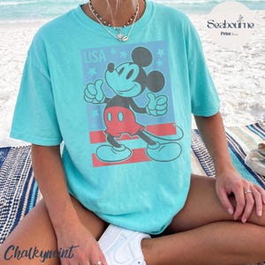 K&ouml;nnte beinhalten: Ein hellt&uuml;rkisfarbenes T-Shirt mit kurzen &Auml;rmeln und einem Grafikdruck von Mickey Mouse, der einen Daumen hoch zeigt. Das Design enth&auml;lt den Text USA, blaue Sterne und rote horizontale Streifen. Das Shirt hat einen Rundhalsausschnitt.