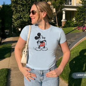 May include: Light blue t-shirt featuring a classic Mickey Mouse graphic. The cartoon character is depicted in black, red, and white. The shirt is a crew neck style and is paired with blue jeans. The words "LIGHT BLUE" are in the bottom right corner.