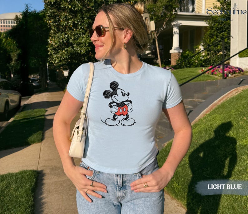 May include: Light blue t-shirt featuring a classic Mickey Mouse graphic. The cartoon character is depicted in black, red, and white. The shirt is a crew neck style and is paired with blue jeans. The words "LIGHT BLUE" are in the bottom right corner.