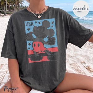 K&ouml;nnte beinhalten: Ein anthrazitfarbenes Kurzarm-T-Shirt mit einer Grafik von Mickey Mouse, die einen Daumen nach oben zeigt. Das Design enth&auml;lt ein blaues Rechteck mit dem Text USA, schwarzen Sternen und roten horizontalen Streifen auf der Vorderseite.
