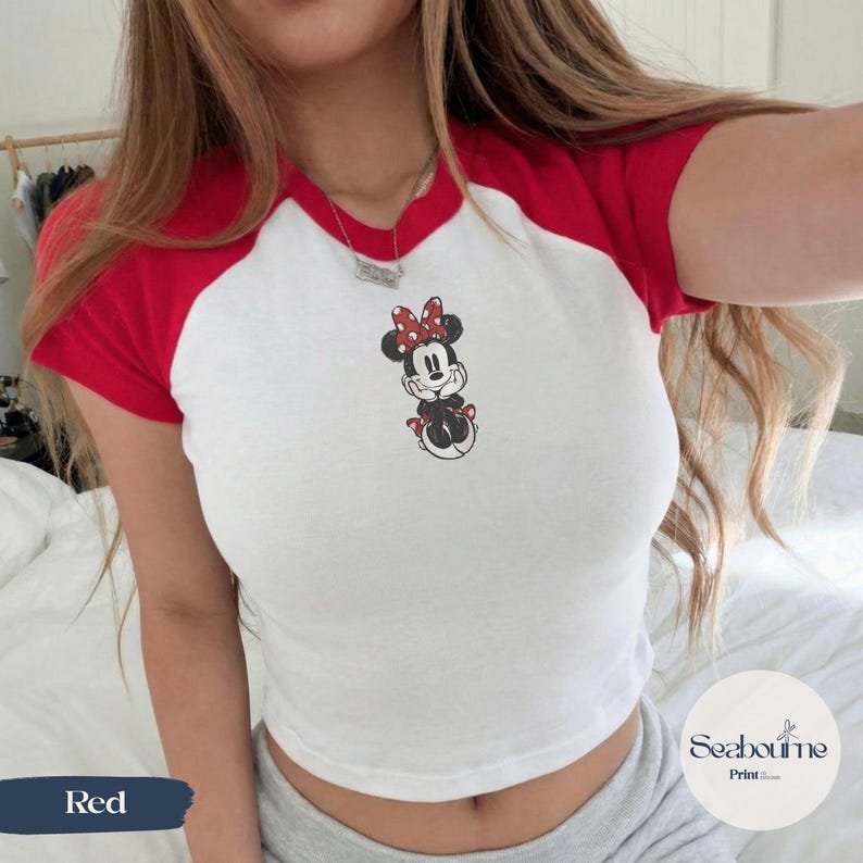 May include: White crop top with red short sleeves and collar featuring a Minnie Mouse graphic. The cartoon character is wearing a red bow with white polka dots. The word "Red" is printed on a blue rectangle.