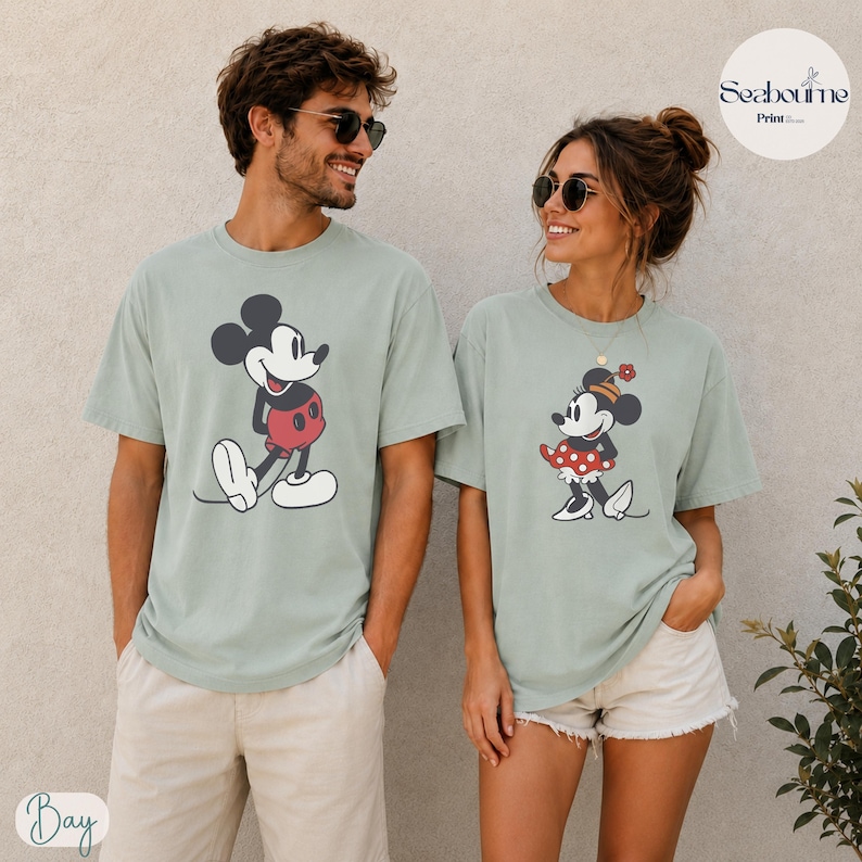 May include: Two sage green t-shirts featuring Mickey and Minnie Mouse. Mickey is wearing red shorts, and Minnie is wearing a red polka dot skirt. The shirts are part of a matching set, perfect for couples or friends.