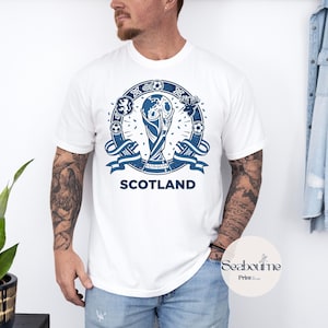 May include: White t-shirt featuring a blue graphic design with the word "SCOTLAND" at the bottom. The design includes a trophy, soccer balls, and a circular emblem. The shirt is made of a soft material.