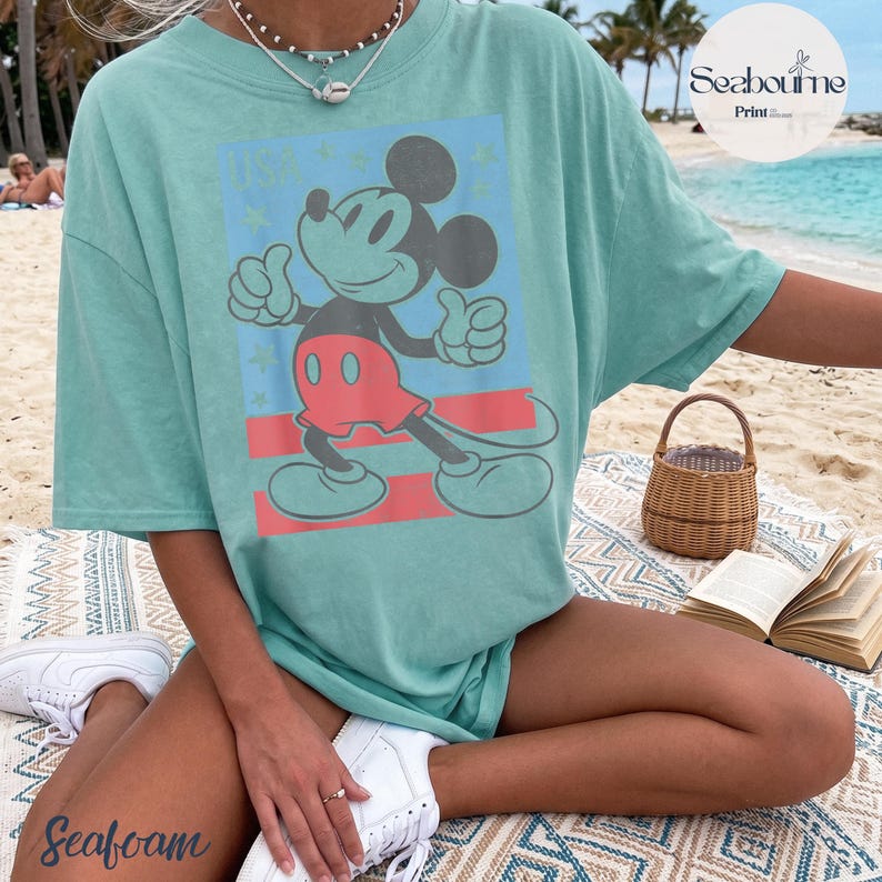 K&ouml;nnte beinhalten: Ein meergr&uuml;nes Kurzarm-T-Shirt mit einer Grafik von Mickey Mouse, der den Daumen hochh&auml;lt. Das Design enth&auml;lt den Text USA, rote horizontale Streifen und kleine Sternakzente. Das T-Shirt hat einen Rundhalsausschnitt und eine lockere Passform.