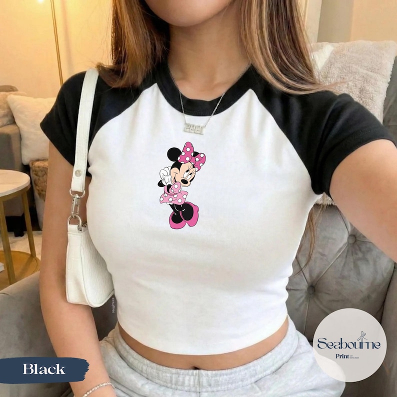 May include: White and black raglan crop top featuring a Minnie Mouse graphic. The graphic shows Minnie Mouse in a pink polka dot dress and bow. The top has black sleeves and a white body. The word "Black" is visible on the bottom left.