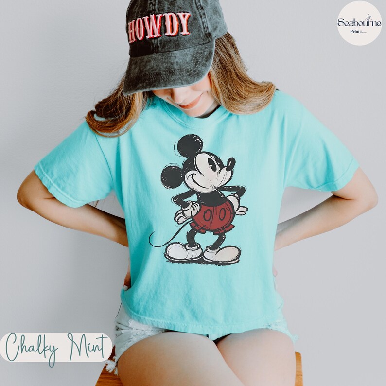 May include: A light blue t-shirt featuring a classic Mickey Mouse graphic. The cartoon character is depicted in black, red, and white. The shirt is a Chalky Mint color and is paired with a black cap with the word "HOWDY".