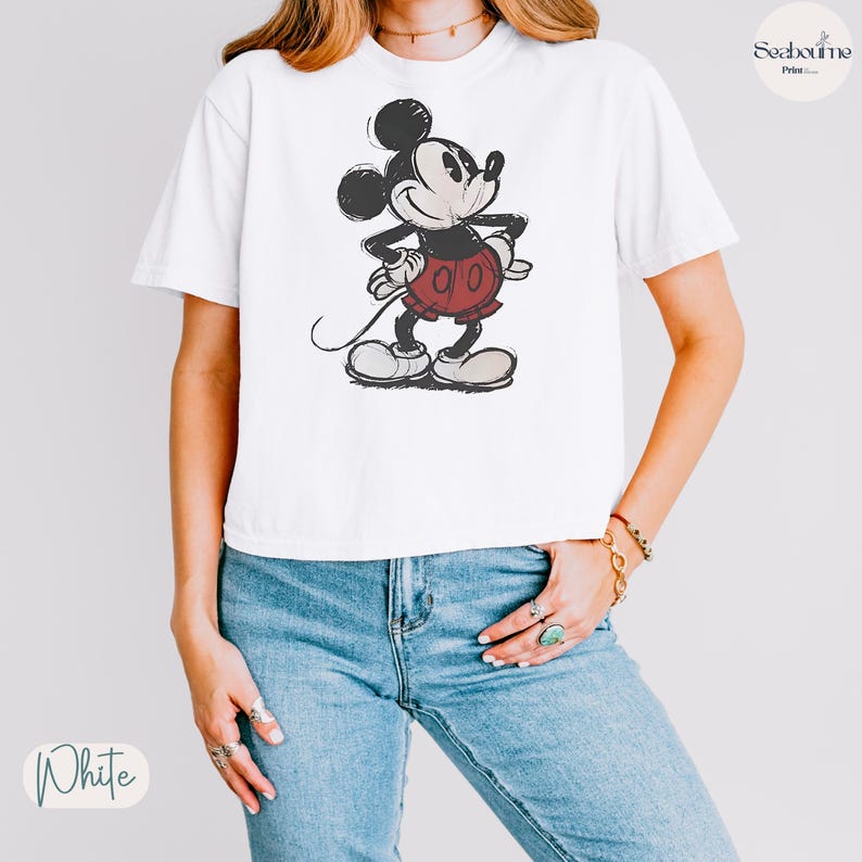 May include: White t-shirt featuring a distressed graphic of Mickey Mouse in a classic pose. Mickey is depicted in black, red, and beige. The shirt is paired with blue jeans. The word "White" is printed on the bottom left.