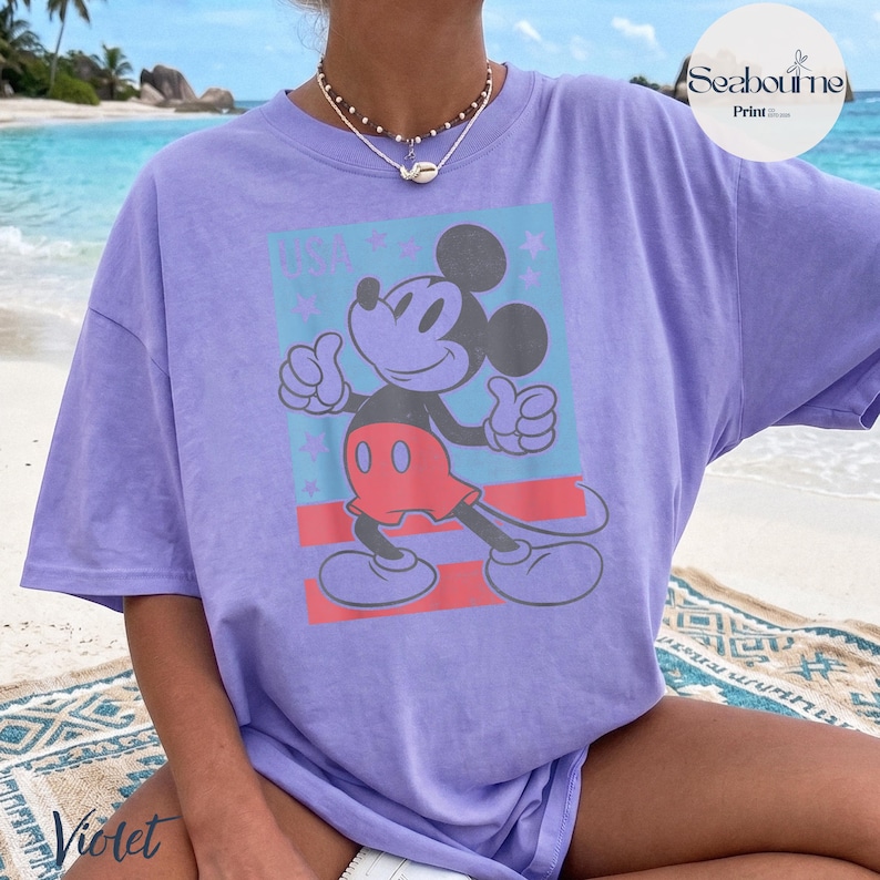 K&ouml;nnte beinhalten: Ein hellviolettes T-Shirt mit einer Grafik von Mickey Mouse, der beide Daumen nach oben zeigt. Das Design enth&auml;lt einen hellblauen rechteckigen Hintergrund mit roten Streifen und kleinen Sternen sowie den Schriftzug USA in abgenutzter Schrift.