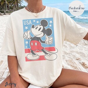 K&ouml;nnte beinhalten: Ein elfenbeinfarbenes Kurzarm-T-Shirt mit einer Grafik von Mickey Mouse, der einen Daumen hoch zeigt. Das Design enth&auml;lt den Text USA, wei&szlig;e Sterne auf blauem Grund und rote horizontale Streifen auf der Vorderseite des Kleidungsst&uuml;cks.