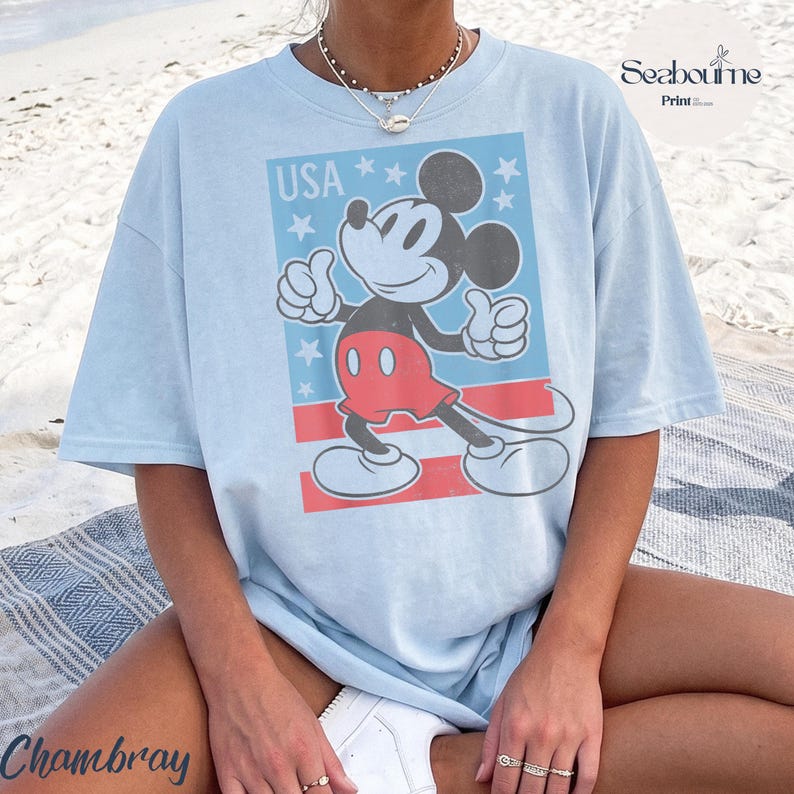 K&ouml;nnte beinhalten: Ein hellblaues T-Shirt mit einer Grafik von Mickey Mouse in roten Shorts und wei&szlig;en Handschuhen. Das Design enth&auml;lt den Text USA, wei&szlig;e Sterne und rote Streifen. Das T-Shirt hat einen Rundhalsausschnitt und kurze &Auml;rmel mit einem Druck.