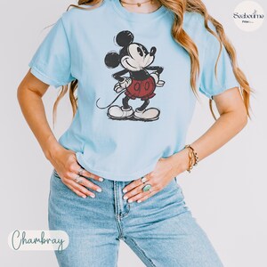May include: Light blue t-shirt featuring a distressed graphic of Mickey Mouse in black, red, and white. The shirt has short sleeves and a crew neck. The word "Chambray" is written in cursive at the bottom.