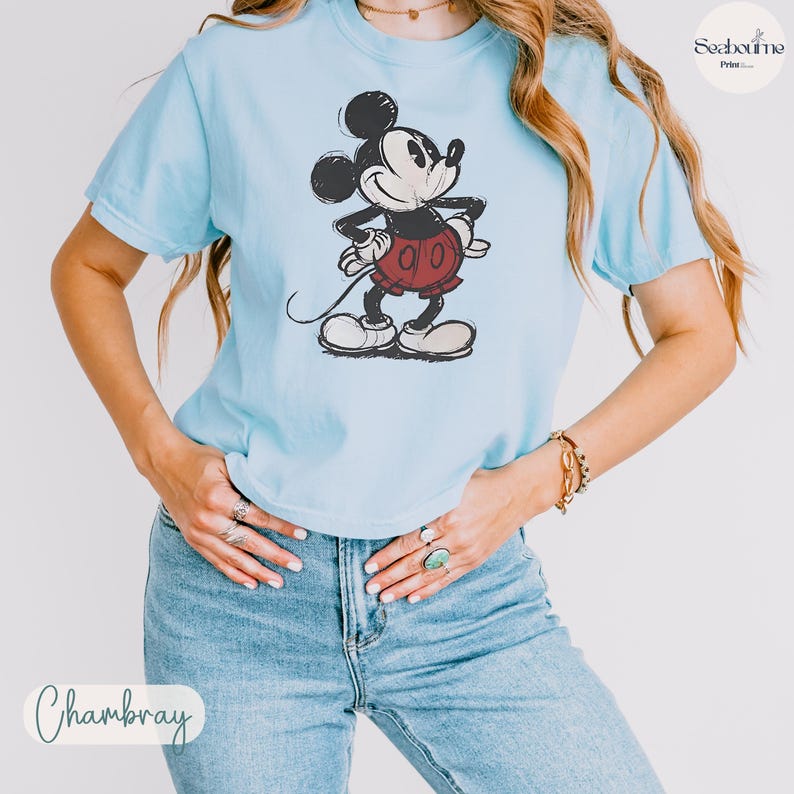 May include: Light blue t-shirt featuring a distressed graphic of Mickey Mouse in black, red, and white. The shirt has short sleeves and a crew neck. The word "Chambray" is written in cursive at the bottom.