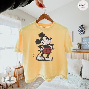 May include: Yellow t-shirt featuring a classic Mickey Mouse graphic. The cartoon character is depicted in black, white, red, and yellow. The shirt is hanging on a wooden hanger.