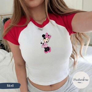 May include: White and red raglan-style crop top featuring a Minnie Mouse graphic. The graphic shows Minnie Mouse in a pink polka-dot dress and bow. The top has red sleeves and a red neckline.