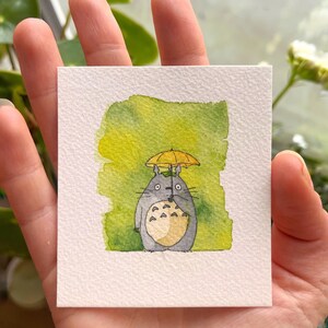 May include: A watercolour painting of a grey Totoro character holding a black umbrella, with a yellow umbrella over its head. The background is a blend of green and yellow watercolour paint. The artwork is on a square, textured, white paper.