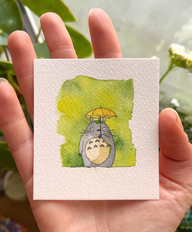 May include: A watercolour painting of a grey Totoro character holding a black umbrella, with a yellow umbrella over its head. The background is a blend of green and yellow watercolour paint. The artwork is on a square, textured, white paper.