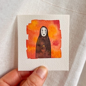 Op de afbeelding: Waterverfschilderij van een personage uit Spirited Away, de No-Face-geest, in een donkerbruine jurk met een wit maskerachtig gezicht. De achtergrond is een mix van oranje en rode tinten, waardoor een warme, levendige achtergrond ontstaat. Het kunstwerk staat op een klein vierkant papier.