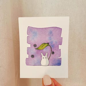 Puede incluir: Ilustración en acuarela de una criatura blanca con forma de conejo y ojos grandes, de pie bajo una hoja verde. El fondo es un lavado de acuarela púrpura y azul con tres pequeñas criaturas negras y borrosas.
