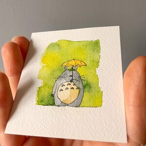 May include: Watercolour painting of a grey Totoro character with a yellow umbrella. The artwork is on a white square card with a green and yellow watercolour background. The character has a cream-coloured belly and black whiskers.