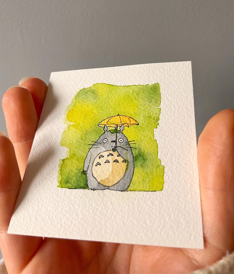 May include: Watercolour painting of a grey Totoro character with a yellow umbrella. The artwork is on a white square card with a green and yellow watercolour background. The character has a cream-coloured belly and black whiskers.