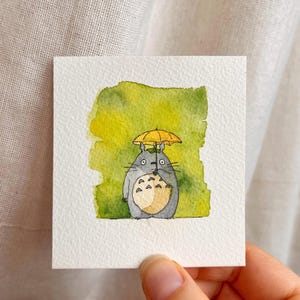 May include: Watercolour painting of a grey Totoro character holding a yellow umbrella. The character has a white belly and is set against a green and yellow watercolour background. The artwork is on a square white paper.