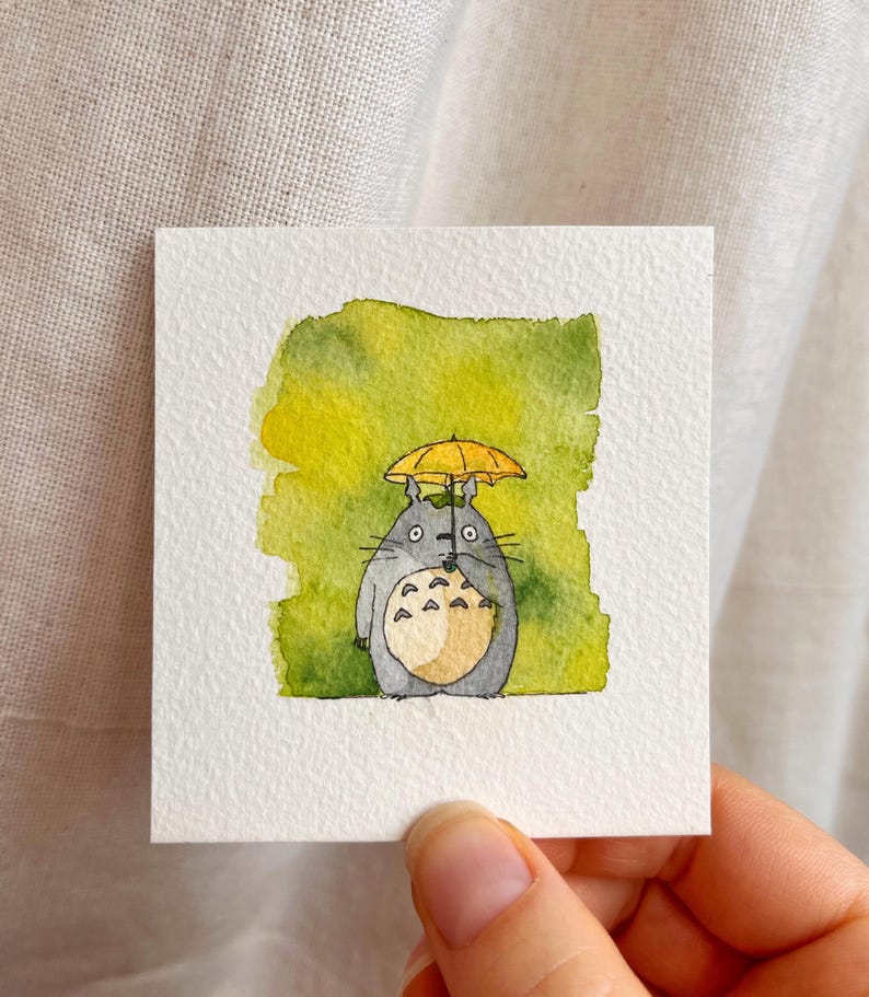 May include: Watercolour painting of a grey Totoro character holding a yellow umbrella. The character has a white belly and is set against a green and yellow watercolour background. The artwork is on a square white paper.