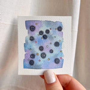 May include: A watercolour painting depicting a fantasy scene with black, round characters with eyes and mouths, set against a blue, purple, and pink background. Small, colourful stars are scattered throughout. The artwork is on a square piece of paper.