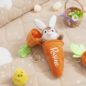 May include: A plush carrot toy in orange, with the word "Rasine" in white, containing a brown and white bunny. A small yellow chick and a miniature basket with flowers are also visible. The items are on a beige dotted surface.