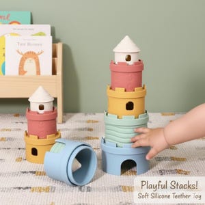 以下が含まれることがあります： お城の塔の形をした、カラフルなシリコン製の積み重ねおもちゃ。おもちゃは、水色のベース、黄色のミドルセクション、ピンクのトップ、白い屋根が特徴です。「Playful Stacks! Soft Silicone Teether Toy」というテキストが表示されています。