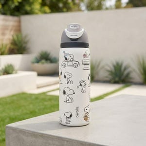 Puede incluir: Una botella de agua Owala blanca con ilustraciones en blanco y negro del personaje Snoopy. La botella tiene una tapa gris con un mecanismo de bot&oacute;n y el texto "Lift to Unlock". La botella est&aacute; sobre una superficie de hormig&oacute;n.