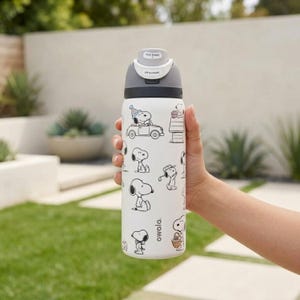 Puede incluir: Una botella de agua Owala blanca con ilustraciones de Snoopy en blanco y negro. La botella tiene una tapa gris con un mecanismo de bot&oacute;n y el texto "Lift to Unlock". La botella est&aacute; sujeta por una persona.