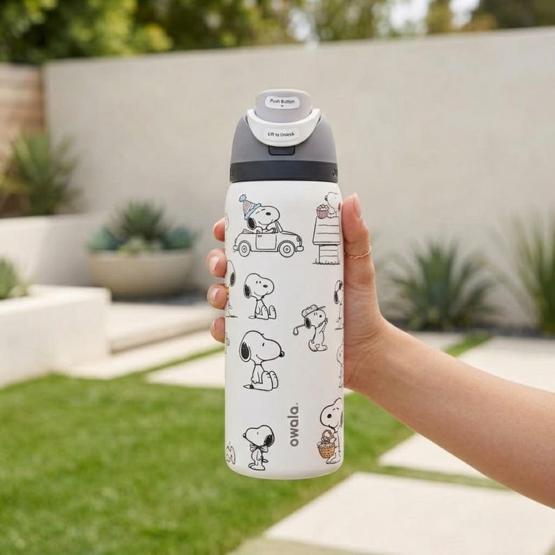 Puede incluir: Una botella de agua Owala blanca con ilustraciones de Snoopy en blanco y negro. La botella tiene una tapa gris con un mecanismo de bot&oacute;n y el texto "Lift to Unlock". La botella est&aacute; sujeta por una persona.