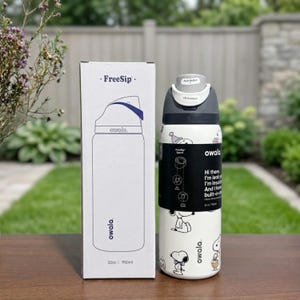 Puede incluir: Una botella de agua Owala FreeSip blanca con un dise&ntilde;o de Snoopy en blanco y negro, junto a una caja a juego. La botella tiene una tapa gris con un bot&oacute;n y una banda negra con texto. La caja dice "FreeSip" y "owala."