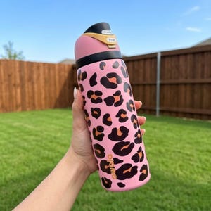 Peut inclure: Bouteille d'eau rose avec un motif léopard noir et orange. La bouteille a un couvercle noir avec un bouton-poussoir et un accent doré. La marque "owala" est imprimée sur le côté.