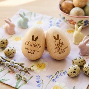 Peut inclure: Deux œufs de Pâques en bois avec des oreilles de lapin gravées et les noms "Charlotte" et "Isla", avec "First Easter 2025" et "Happy Easter 2025". Ils sont posés sur un tissu à motifs floraux avec des lapins en céramique, des œufs tachetés et un bol d'œufs.
