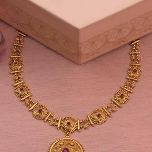 May include: Gold-toned necklace with a large, circular pendant and smaller circular accents, each featuring a red gemstone. The necklace has an ornate, detailed design and is displayed on a pink surface.