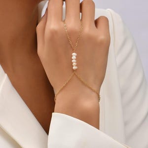 Könnte beinhalten: Eine goldfarbene Handkette mit filigranem Kettendesign. Die Kette verbindet sich mit einem Armband und erstreckt sich bis zu einem Fingerring, wobei eine Reihe von fünf weißen Perlen die Mitte ziert. Der Schmuck wird an einer Hand getragen, mit einem weißen Blazer im Hintergrund.