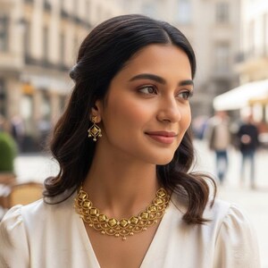 May include: A gold-coloured necklace and matching earrings are worn by a woman. The necklace has a repeating geometric pattern with small pearl details. The earrings have a similar design. The woman is wearing a white dress.