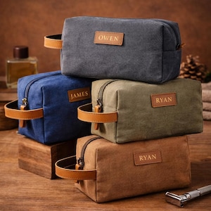 Könnte beinhalten: Stapel personalisierter Kulturbeutel aus Canvas in verschiedenen Farben: Grau, Blau, Olivgrün und Braun. Jede Tasche hat ein Lederetikett mit einem Namen und einen passenden Ledergriff. Die Taschen stehen auf einer Holzoberfläche.