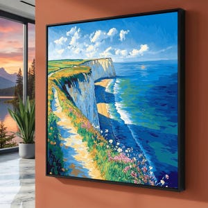 Puede incluir: Una pintura de paisaje enmarcada representa una escena costera con un acantilado blanco, una playa de arena y un océano azul vibrante bajo un cielo brillante con nubes. Un camino serpentea por el borde del acantilado, adornado con flores coloridas. La obra de arte está en un marco negro.