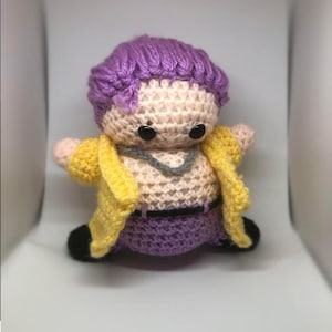 Könnte beinhalten: Handgemachte Häkelpuppe mit lila Perücke, schwarzen Augen und gelber Jacke. Die Puppe hat einen hellen Hautton, eine graue Halskette und eine lila Hose. Die Schuhe der Puppe sind schwarz.