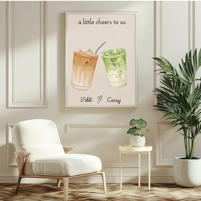 May include: A rectangular wall art print on a light beige background featuring an illustration of an iced coffee and an iced matcha latte. The text reads a little cheers to us at the top and Vikki ♡ Casey at the bottom in black script.