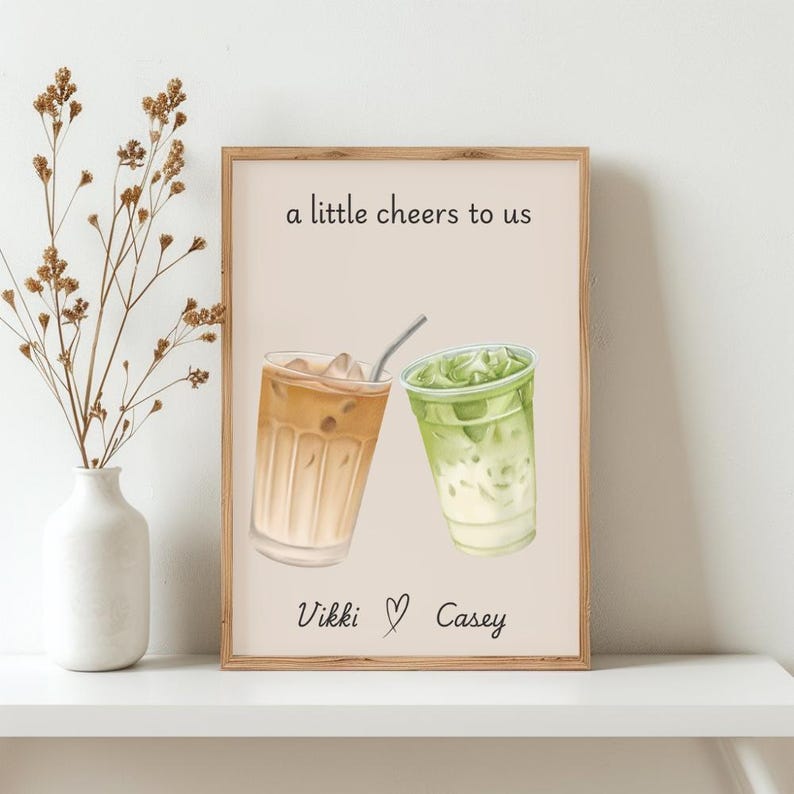 May include: A rectangular wall art print featuring a watercolor illustration of two iced drinks, one brown and one green, with the text "a little cheers to us" at the top and the names "Vikki" and "Casey" separated by a heart symbol at the bottom.