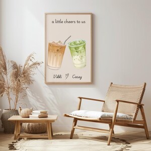 May include: A rectangular wall art print featuring a light beige background with two iced drinks in clear glasses, one tan and one green. The text "a little cheers to us" appears at the top, and "Vikki ♡ Casey" is printed at the bottom in black.
