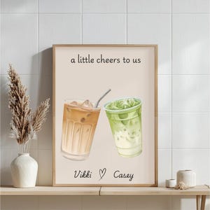 May include: A vertical wall art print on a light beige background featuring an illustration of an iced coffee and an iced matcha latte. The text reads a little cheers to us at the top and Vikki with a heart symbol and Casey at the bottom.