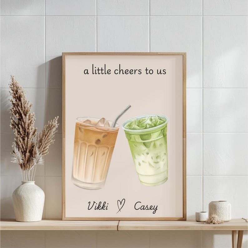 May include: A vertical wall art print on a light beige background featuring an illustration of an iced coffee and an iced matcha latte. The text reads a little cheers to us at the top and Vikki with a heart symbol and Casey at the bottom.