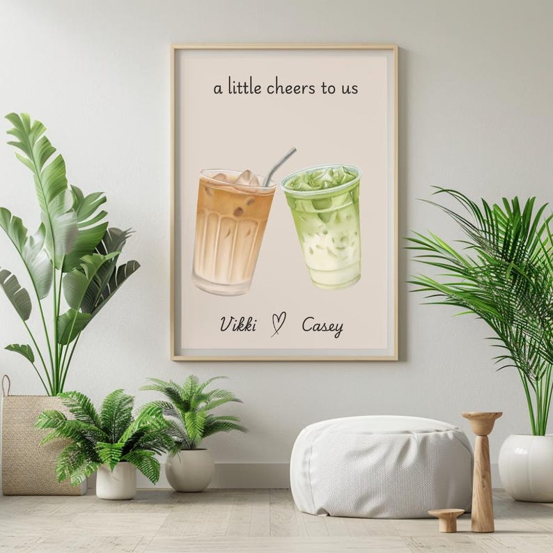 May include: A rectangular wall art print on a light beige background featuring an illustration of two iced drinks, one brown and one green, with a straw. The text reads a little cheers to us above the drinks and Vikki heart Casey below.