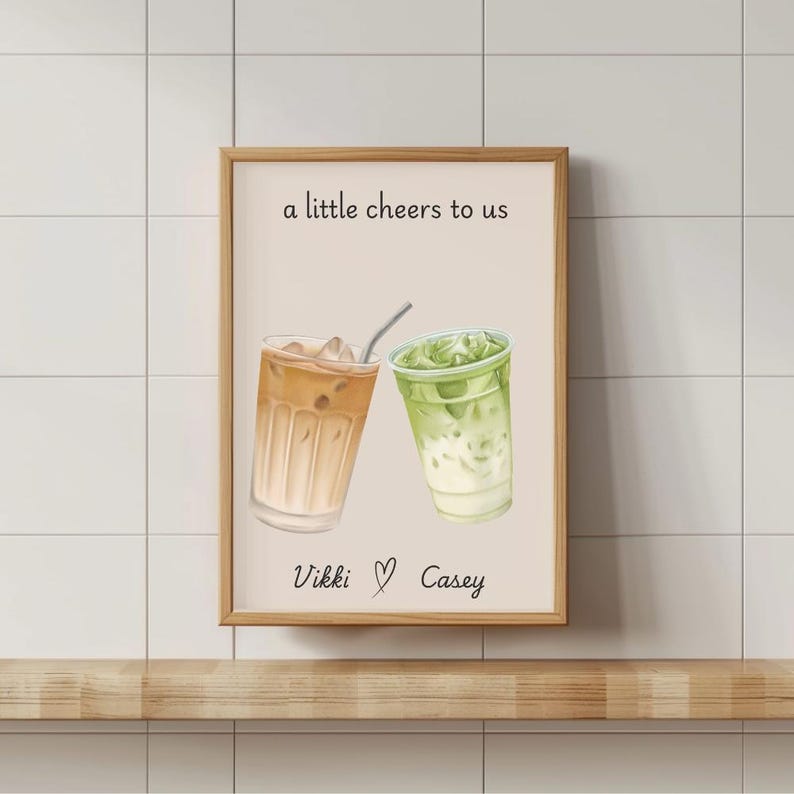 May include: A framed wall art print featuring a light beige background with an illustration of an iced coffee and an iced matcha latte. The text reads a little cheers to us above the drinks and Vikki ♡ Casey below in black script lettering.
