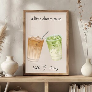 May include: A framed wall art print featuring a watercolor illustration of two iced drinks, one brown and one green, with the text 'a little cheers to us' at the top and the names 'Vikki' and 'Casey' separated by a heart symbol at the bottom.