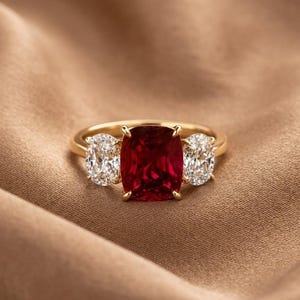May include: A gold ring featuring a large, cushion-cut, deep red gemstone at the centre, flanked by two smaller, oval-cut, clear white gemstones. The ring has a polished gold band and a prong setting that secures each of the three stones in place.